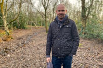 A man in a waxed jacket and jeans stands on a leafy path in some woodland