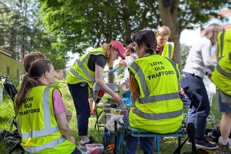 Volunteers from Love Old Trafford sit in the shade of large trees, sharing food on a break. They're wearing high-viz jackets that say "Love Old Trafford" on the back