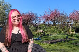 A lady with pink and brown long straight hair wearing a black dress and glasses, in front of a large green garden space