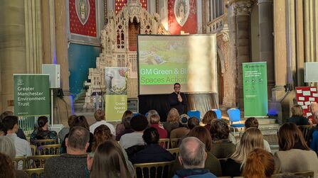 Mayor of Greater Manchester Andy Burnham addresses a large crowd in the main hall at Gorton Monastery. Behind him, a green screen reads 'GM Green & Blue Spaces Action Network'.