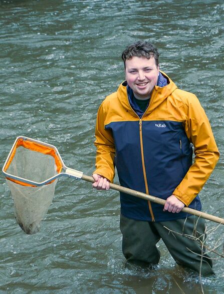 Sam is standing in a body of water holding a net and wearing a blue and yellow coat
