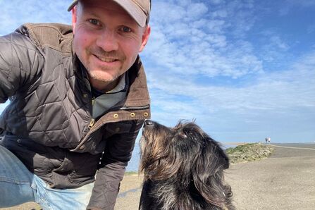 A man in a baseball cap, a waxed jacket and jeans smiles at the camera next to a black dog, on a beach