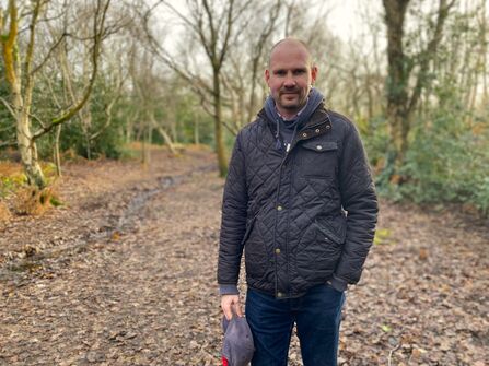 A man in a waxed jacket and jeans stands on a leafy path in some woodland