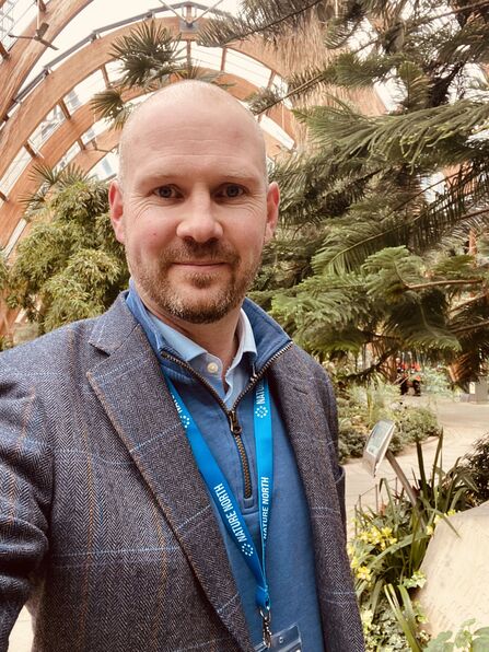 A selfie of a man in a grey suit with a blue lanyard standing in front of pine trees
