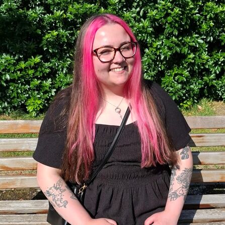 A lady with pink and brown hair wearing glasses and a black dress sits on a bench in front of a background of green bushes